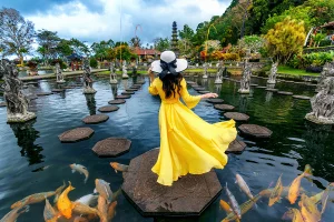 A woman in a vibrant yellow dress standing on a stone pathway at Tirta Gangga Water Palace surrounded by koi fish